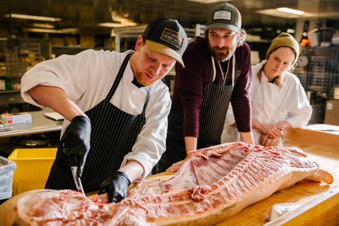 a man butchering meat while two others watch